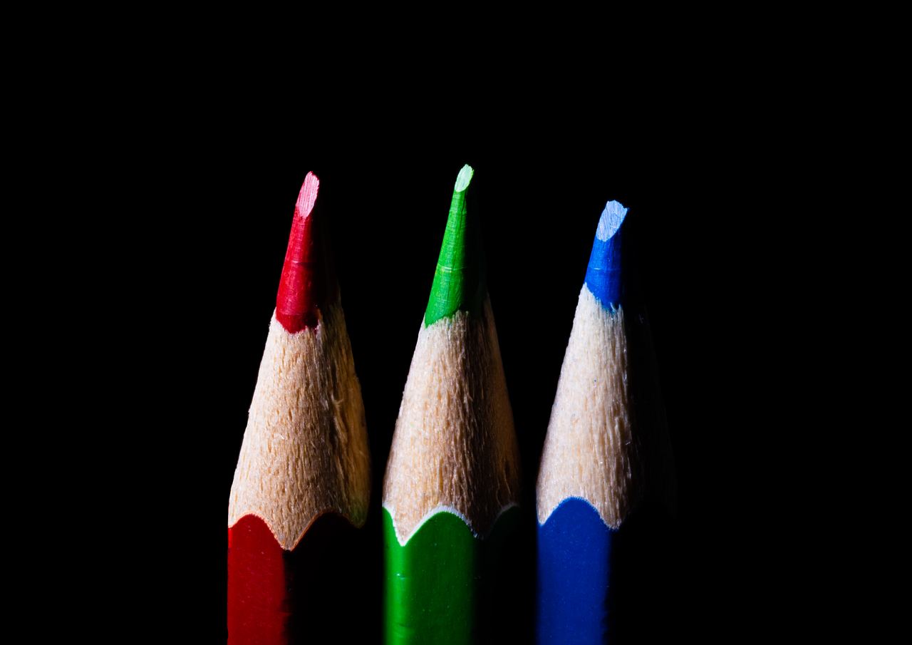 The sharpened tips of three coloured pencils, red, green and blue, standing upright against black, the bare wood glowing under the light.
