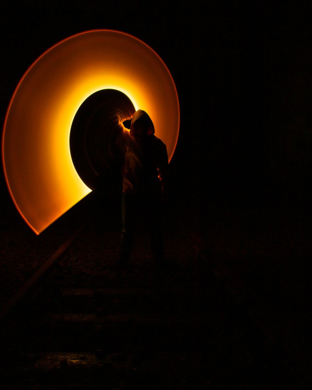 A hooded figure stands silhouetted on railway tracks at the mouth of a tunnel, ringed by a swirling halo of orange light painted during a long exposure.
