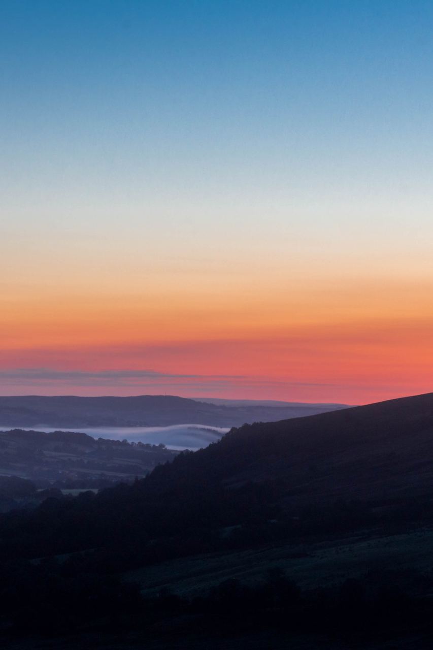 A valley at first light: dark layered hills under a band of mist, the sky burning from coral at the horizon up into clear blue.