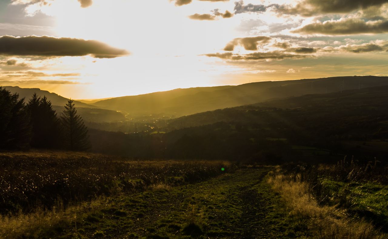 A valley at sunset, golden light flooding between dark hills with a grassy field track leading into the foreground.