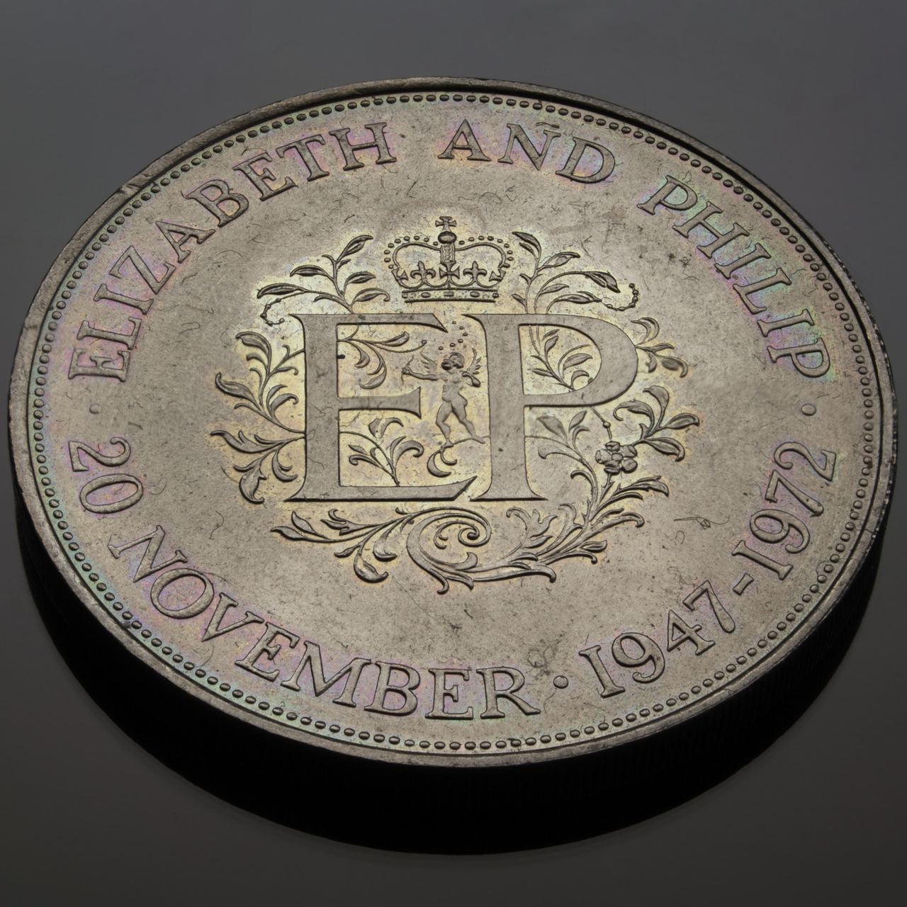 A close up of a 1972 Elizabeth and Philip silver wedding crown, the EP monogram, crown and lettering catching faint rainbow toning.