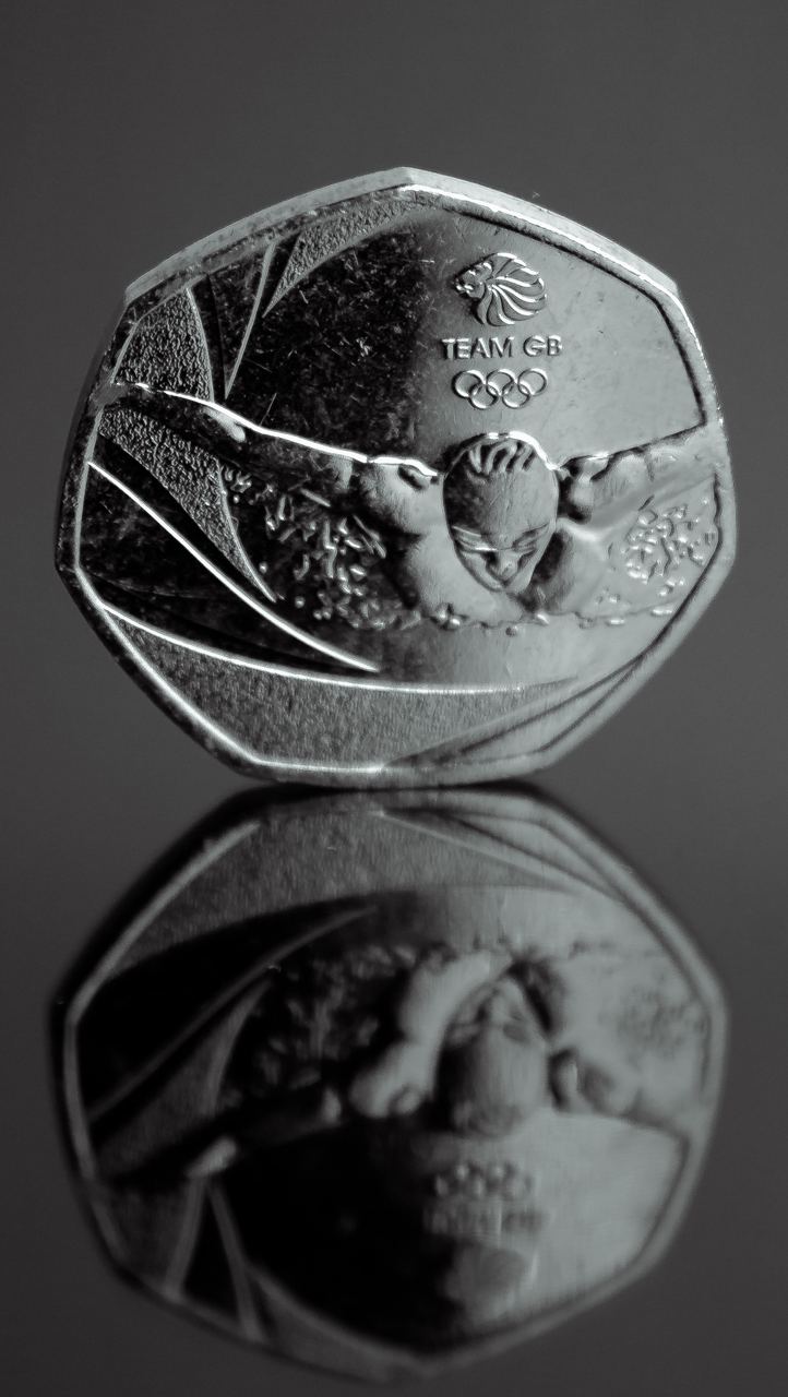 A close up of a Team GB 50p coin showing a swimmer mid stroke, standing upright with its mirrored reflection below.