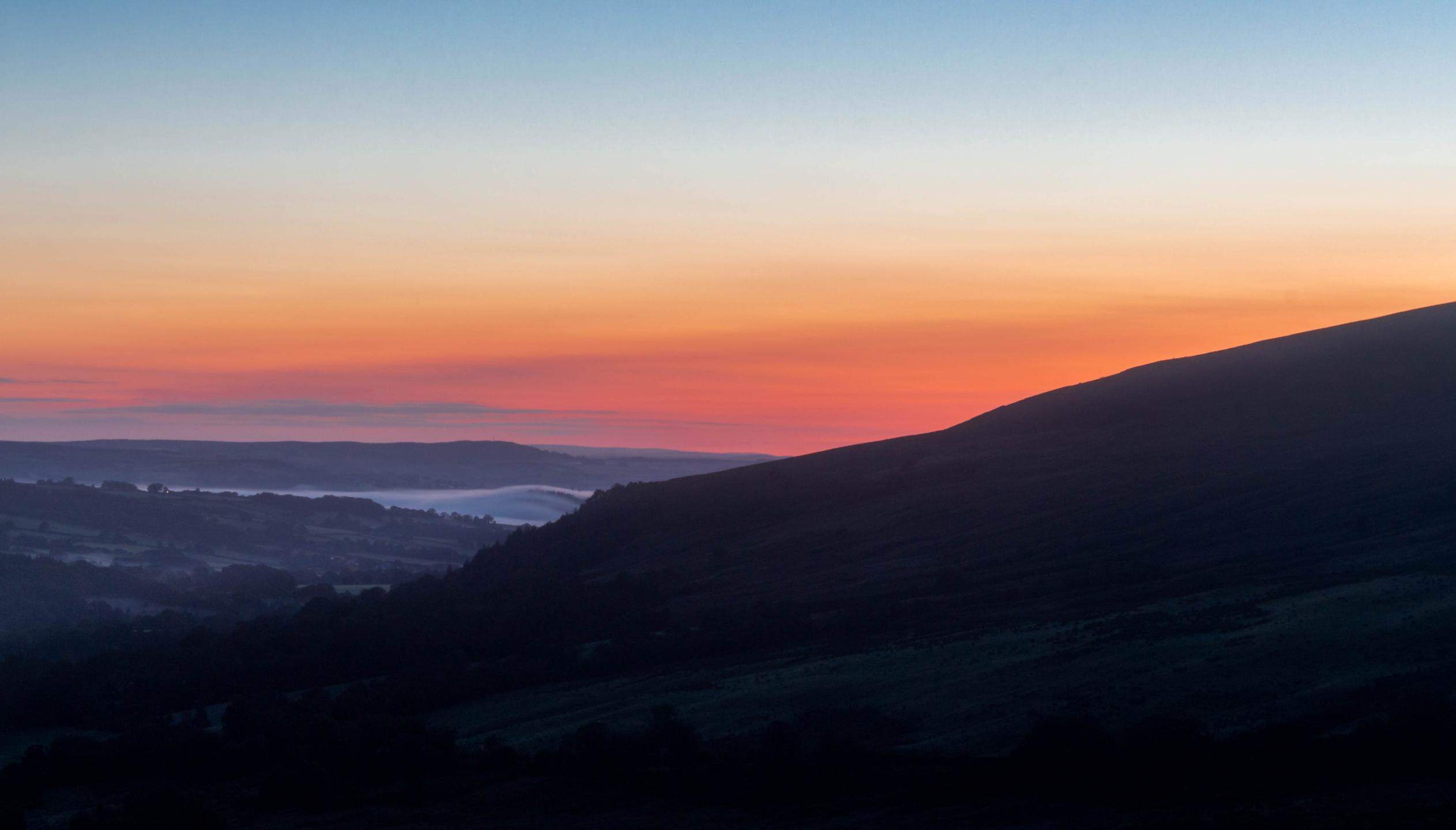 Wide view of a Welsh hillside in dark silhouette on the right, with misty valleys on the left and a gradient sunrise sky from deep blue to warm orange.